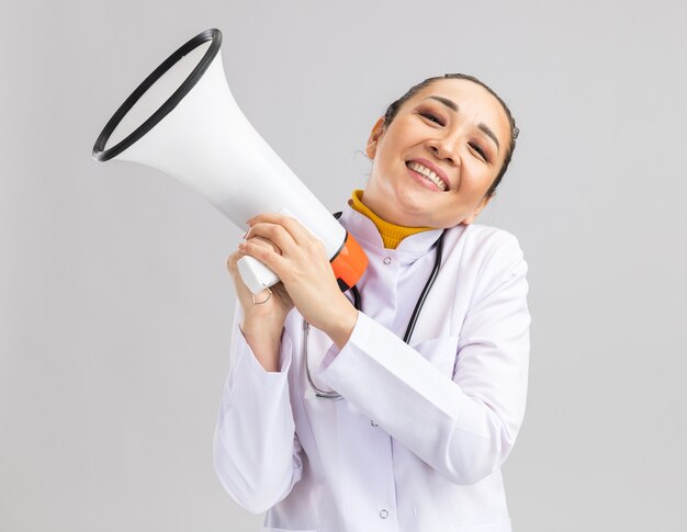 Pleased young woman doctor in white medical coat with stethoscope around neck holding megaphone happy and positive smiling cheerfully standing over white wall