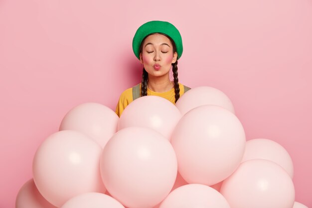 Pleased young woman keeps lips rounded, wears green beret, has eyes closed, has two pigtails, stands near helium balloons