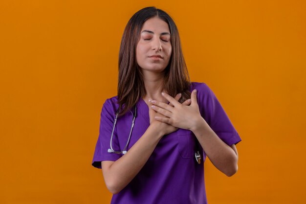 Pleased young woman nurse wearing medical uniform and stethoscope standing with hands crossed on chest gesture feeling positive emotions on isolated orange