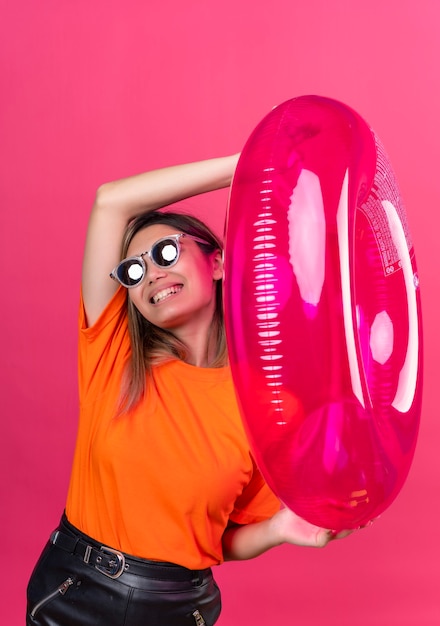 Free photo a pleased young woman in an orange t-shirt wearing sunglasses looking while holding pink inflatable ring on a pink wall