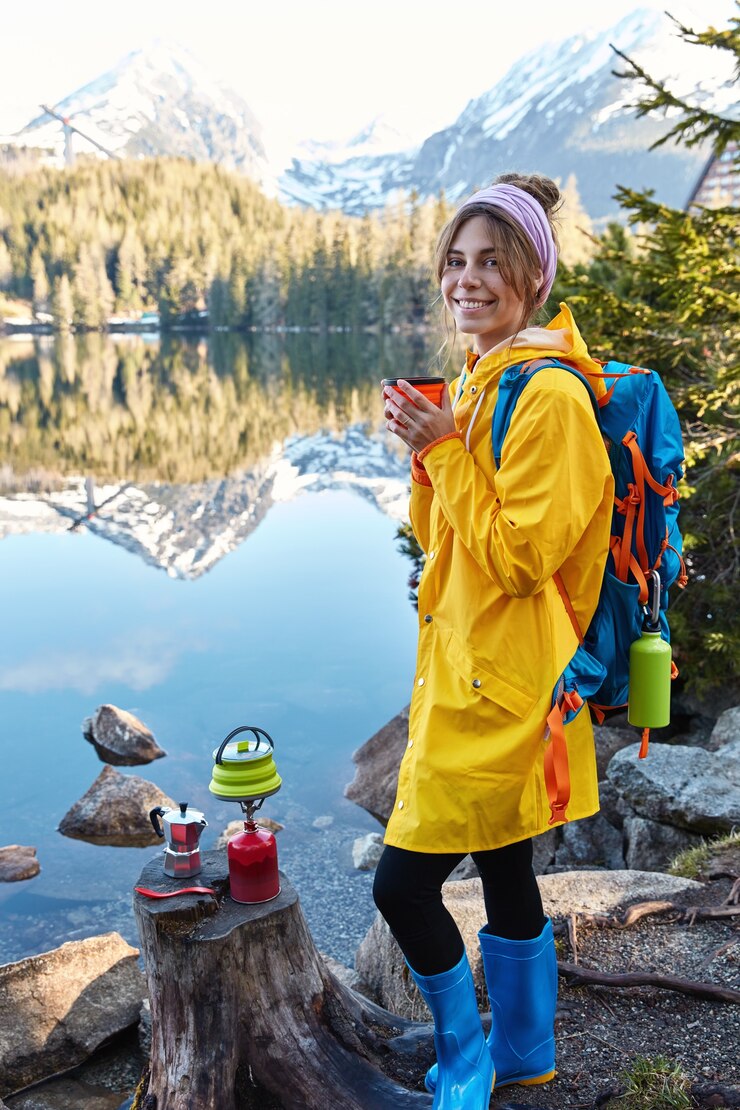 Free Photo pleased young woman smiles gently, drinks hot beverage, wears raincoat and rubber boots, enjoys sunny weather after rain
