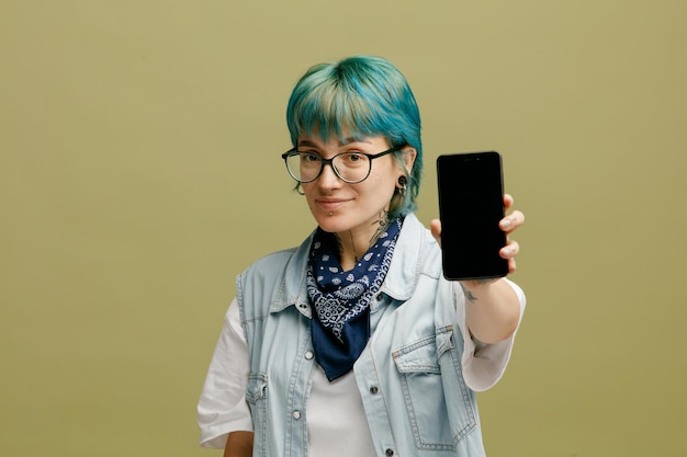 올리브 녹색 배경에 고립 된 휴대 전화를 보여주는 카메라를보고 목에 안경 반다나를 착용하는 기쁘게 젊은 여성