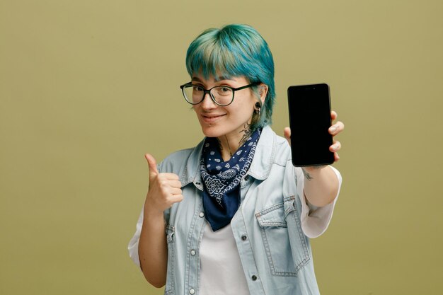 Довольная молодая женщина в очках с банданой на шее смотрит в камеру, протягивая мобильный телефон к камере, показывая большой палец вверх на оливково-зеленом фоне