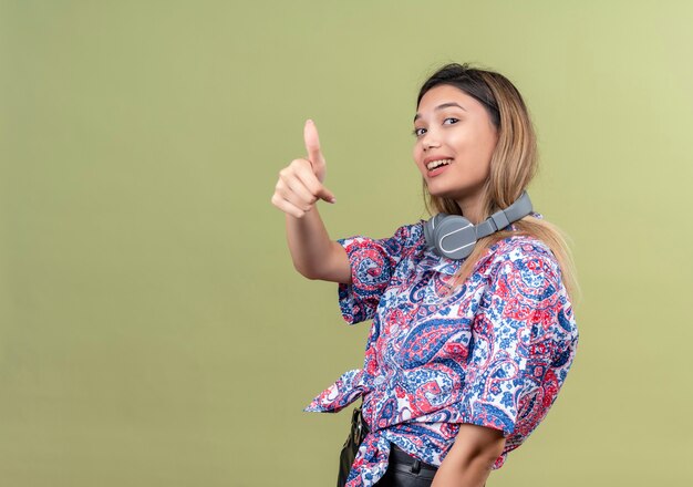 A pleased young woman wearing paisley printed shirt in headphones showing thumbs up