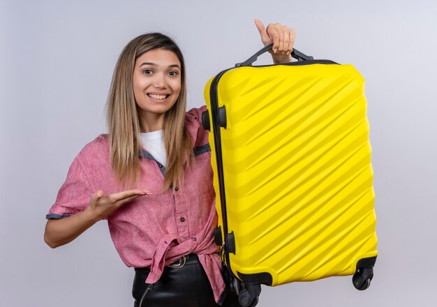 Довольная молодая женщина в красной рубашке показывает свой желтый чемодан, глядя на белую стену