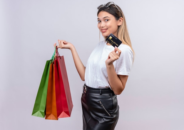 A pleased young woman in white t-shirt wearing sunglasses on her head showing credit card while holding shopping bags on a white wall