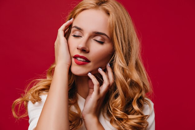 Pleased young woman with blonde hair posing with eyes closed. Carefree fair-haired girl standing on red wall.