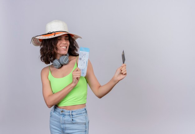 A pleased young woman with short hair in green crop top in headphones wearing sun hat looking at plane tickets and credit card on a white background