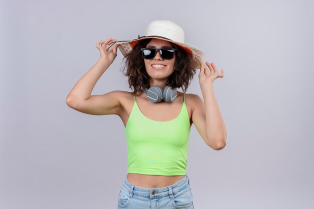 A pleased young woman with short hair in green crop top wearing sunglasses and sun hat on a white background