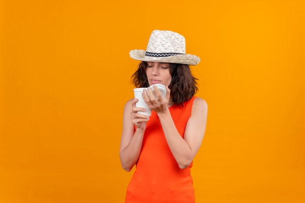 A pleased young woman with short hair in an orange shirt wearing sun hat opening plastic cup and smelling coffee