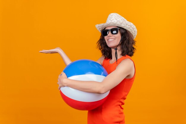膨脹可能なカラフルなボールを保持している太陽の帽子とサングラスを身に着けているオレンジ色のシャツの短い髪の幸せな若い女性