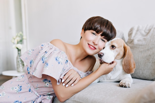 Felice giovane donna con il manicure bianco in posa sognante con il suo cane beagle su grigio chiaro e sorridente
