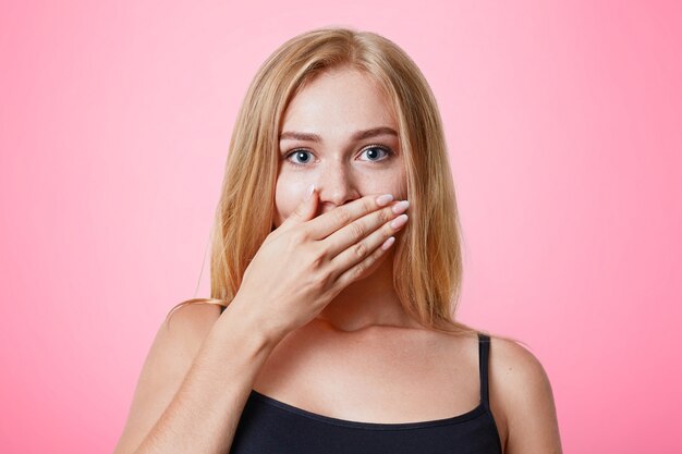 Plesant looking light haired female dressed casually, covers mouth with hand, tries to keep silent and not tell secret, isolated over pink wall. People, beauty, body language concept