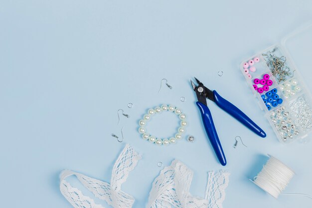 Plier; hook; pearls; lace ribbon; thread spool and plastic beads box on blue background