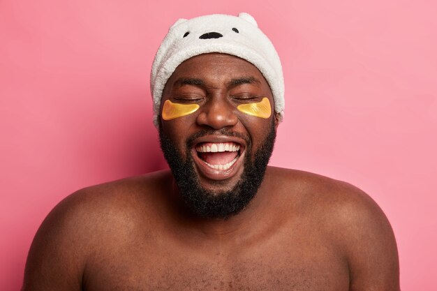 Plump overjoyed dark skinned ethnic man laughs out happily, applies golden patches under lower eyelids, has fun during beauty procedures, wears white hat