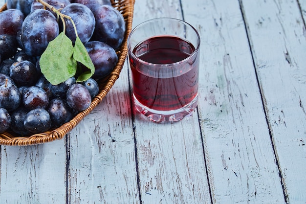 Plums in a basket on blue with a glass of juice.