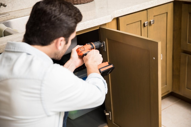 Free photo point of view of a young handyman using a power drill to fix a door in a kitchen cabinet