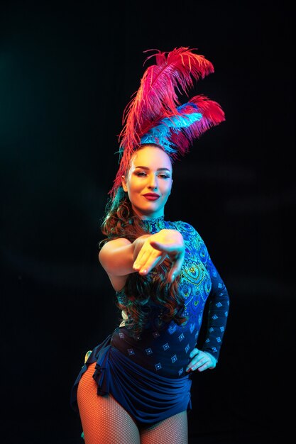 Pointing. Beautiful young woman in carnival, stylish masquerade costume with feathers on black background in neon light. Copyspace for ad. Holidays celebration, dancing, fashion. Festive time, party.