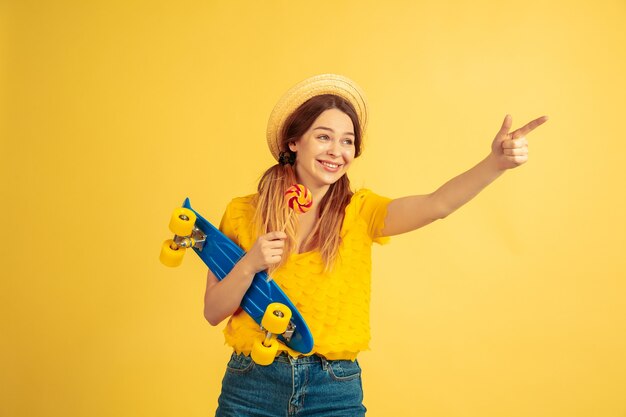 Pointing, calling. Caucasian woman's portrait on yellow studio background. Beautiful female model in hat. Concept of human emotions, facial expression, sales, ad. Summertime, travel, resort.