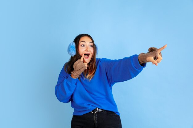Указывая. Портрет кавказской женщины на синем фоне студии. Красивая женская модель в теплой одежде. Понятие человеческих эмоций, выражения лица, продаж, рекламы. Зимнее настроение, Рождество, праздники.