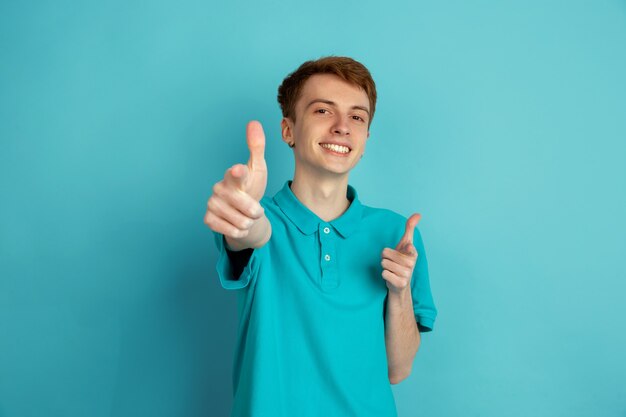 Pointing on, choosing. Caucasian young man's modern portrait isolated on blue  wall, monochrome. Beautiful male model. Concept of human emotions, facial expression, 