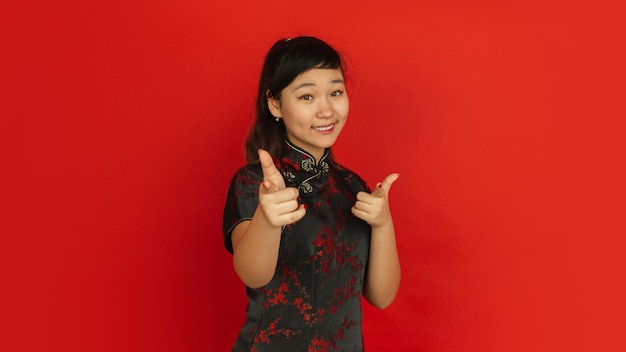 Pointing on, choosing, smiling. Happy Chinese New Year 2020. Asian young girl's portrait on red background. Female model in traditional clothes looks happy. Celebration, human emotions. Copyspace.