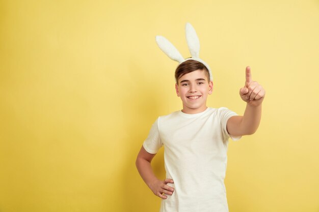 Pointing, choosing you. Caucasian boy as an Easter bunny on yellow studio background. Happy easter greetings. Beautiful male model. Concept of human emotions, facial expression, holidays. Copyspace.