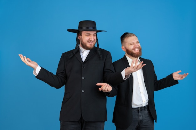 Pointing, greeting. Portrait of a young orthodox jewish men isolated on blue  wall. Purim, business, festival, holiday, celebration Pesach or Passover, judaism, religion concept.