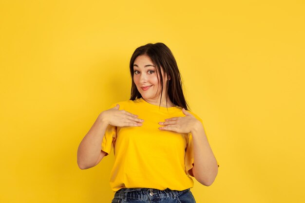 Pointing on herself. Caucasian woman's portrait isolated on yellow  wall. Beautiful female brunette model in casual. Concept of human emotions, facial expression, sales, copyspace.