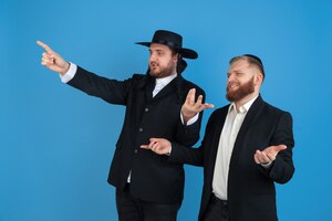 Pointing, inviting. portrait of a young orthodox jewish men isolated on blue wall. purim, business, festival, holiday, celebration pesach or passover, judaism, religion concept.