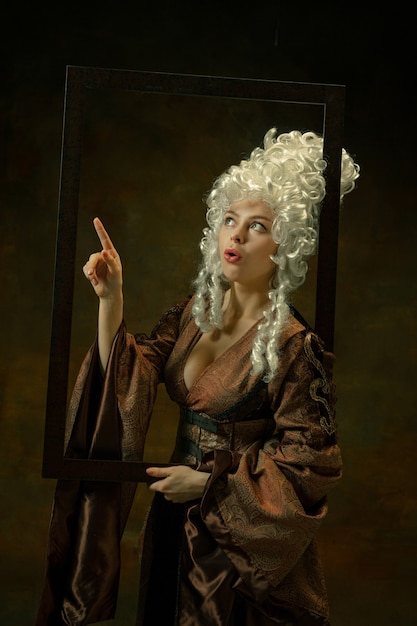 Pointing. Portrait of medieval young woman in vintage clothing with wooden frame on dark background. Female model as a duchess, royal person. Concept of comparison of eras, modern, fashion, beauty.