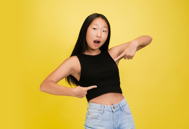 Pointing. portrait of young asian girl isolated on yellow studio background. stylish, trendy. beautiful model, young and emotional. human emotions, facial expression, sales, ad concept. flyer