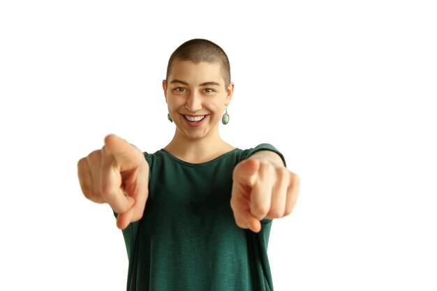 Pointing on. Portrait of young caucasian woman with freaky appearance on white wall. Unusual look with tattoos and bald. Human emotions, facial expression,sales, ad concept. Youth culture.