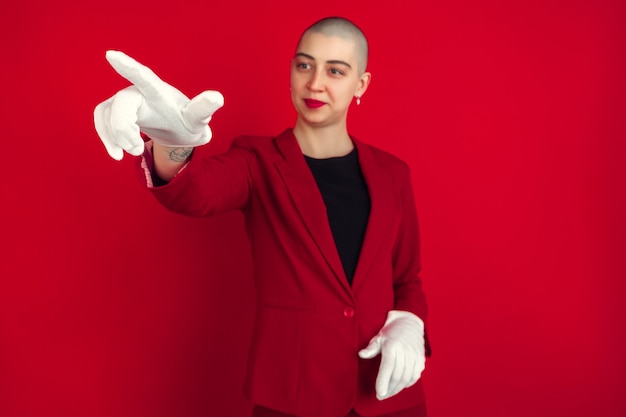 Pointing, showing. Portrait of young caucasian bald woman isolated on red studio wall.