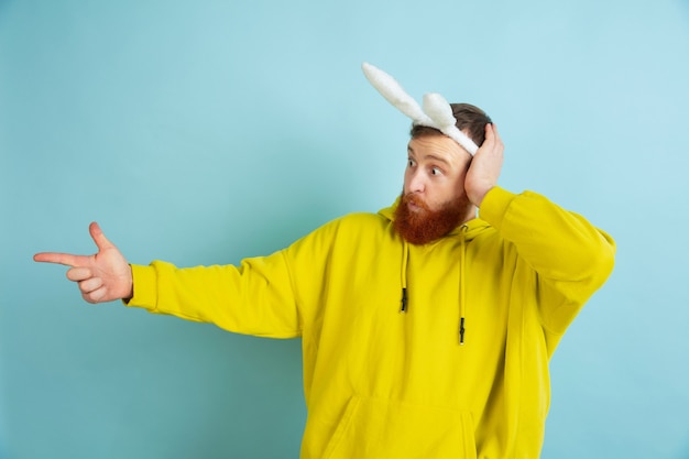 Pointing at side. Caucasian man as an Easter bunny with bright casual clothes on blue studio background.