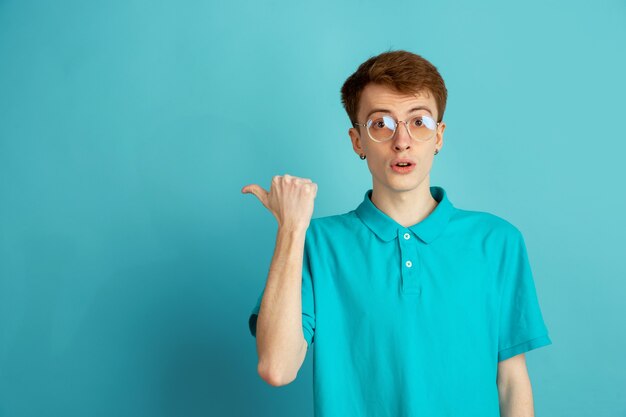 Pointing at side. Caucasian young man's modern portrait on blue studio