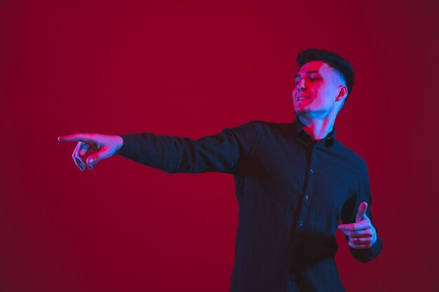 Pointing at side. Caucasian young man's portrait isolated on red  wall in neon light. Beautiful male model in black. Concept of human emotions, facial expression,, youth culture.