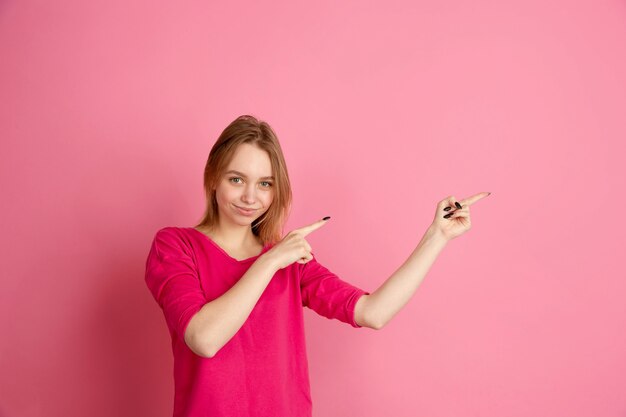 横を指しています。ピンクの壁、モノクロで隔離の白人の若い女性の肖像画。美しい女性モデル。人間の感情、顔の表情、販売、広告、トレンディの概念。