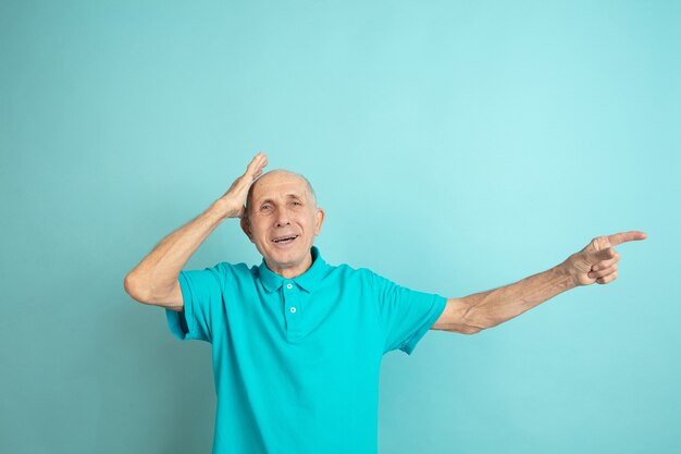 Pointing at side, shocked. Caucasian senior man's portrait on blue studio.