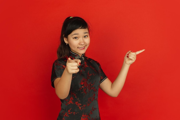 Pointing on and at side, smiling. Happy Chinese New Year. Asian young girl's portrait on red background. Female model in traditional clothes looks happy. Celebration, human emotions. Copyspace.