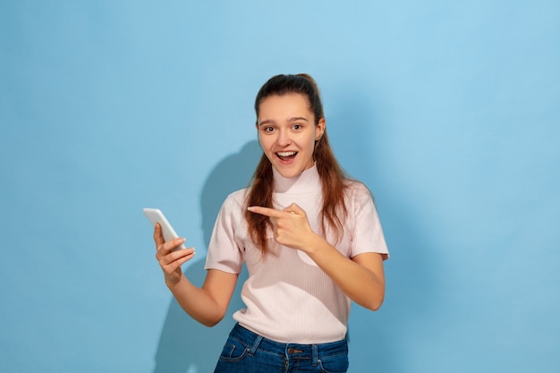 Pointing at smartphone, smiling. Caucasian teen girl's portrait on blue background. Beautiful model in casual wear. Concept of human emotions, facial expression, sales, ad. Copyspace. Looks happy.