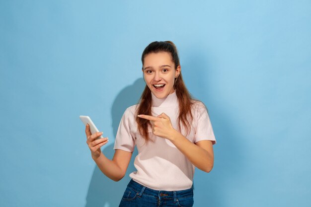 스마트 폰 가리키는, 웃고. 파란색 배경에 백인 십 대 소녀의 초상화입니다. 캐주얼웨어의 아름다운 모델. 인간의 감정, 표정, 판매, 광고의 개념. Copyspace. 행복해 보인다.