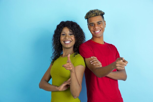 指さし、笑顔。青の背景にカラフルな服を着た若い感情的なアフリカ系アメリカ人の男性と女性。美しいカップル。