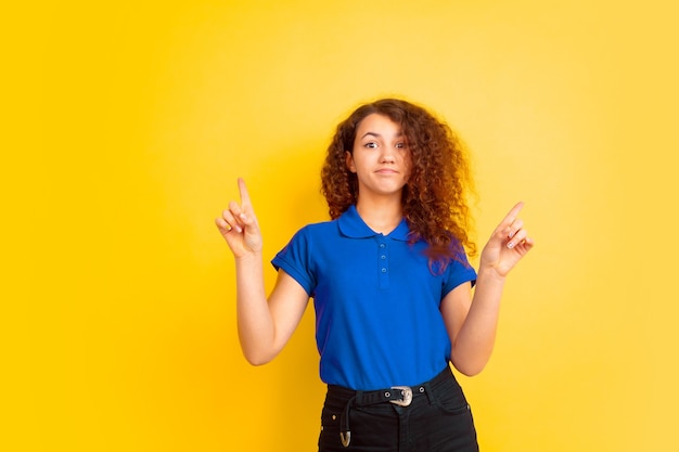 Pointing up, cute. Caucasian teen's girl portrait on yellow studio background. Beautiful female curly model in shirt. Concept of human emotions, facial expression, sales, ad, education. Copyspace.