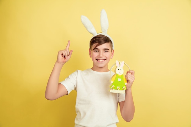 Pointing up. Decorating. Caucasian boy as an Easter bunny on yellow studio background. Happy easter greetings. Beautiful male model. Concept of human emotions, facial expression, holidays. Copyspace.