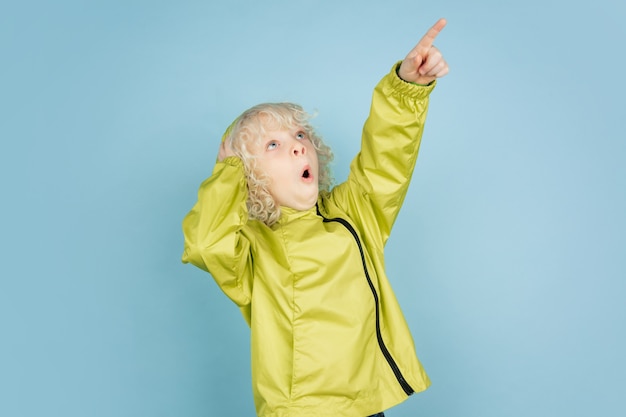 Free photo pointing up. portrait of beautiful caucasian little boy isolated on blue  wall. blonde curly male model. concept of facial expression, human emotions, childhood,  copyspace.