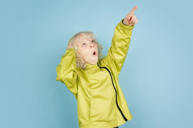 Pointing up. Portrait of beautiful caucasian little boy isolated on blue  wall. Blonde curly male model. Concept of facial expression, human emotions, childhood,  Copyspace.