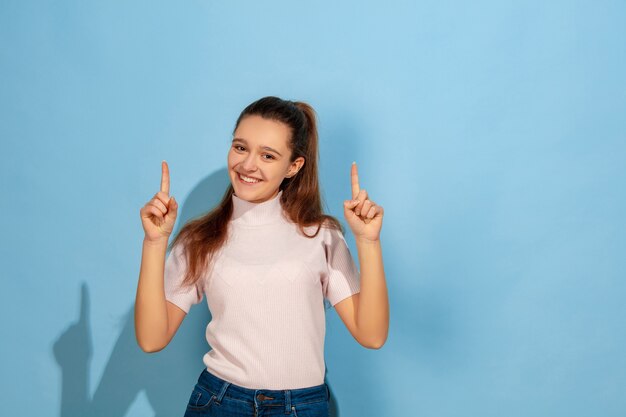 Pointing up, smiling. Caucasian teen girl's portrait on blue background. Beautiful model in casual wear. Concept of human emotions, facial expression, sales, ad. Copyspace. Looks cute, astonished.