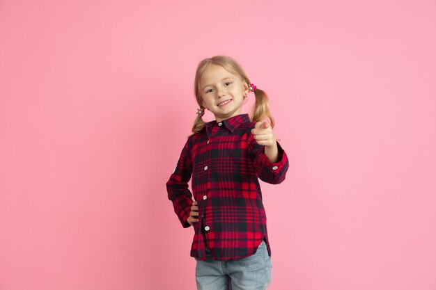 Pointing you, choosing. Caucasian little girl's portrait on pink wall. Beautiful female model with blonde hair. Concept of human emotions, facial expression, sales, ad, youth, childhood.