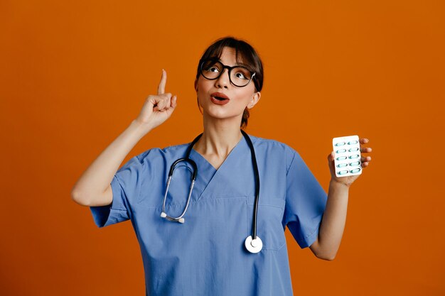 주황색 배경에 격리된 균일한 청진기를 입고 약을 들고 있는 젊은 여성 의사